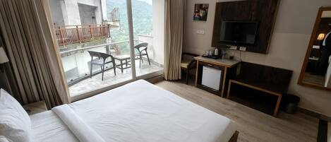 Quarto de hotel com uma cama grande, uma mesa de trabalho com televisão, varanda com cadeiras e vista para as montanhas.