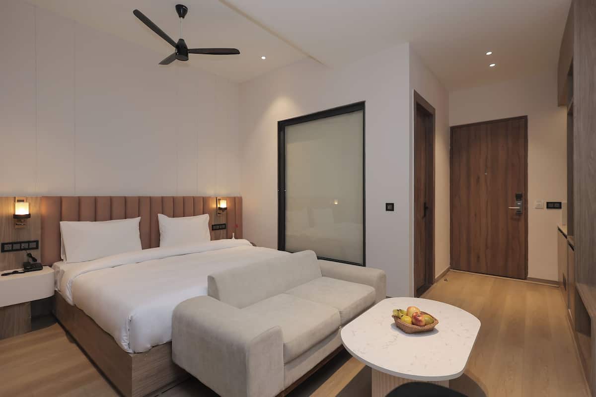 A modern hotel room with a large bed, a sofa, a small table with fruit, a ceiling fan, and a large sliding door.