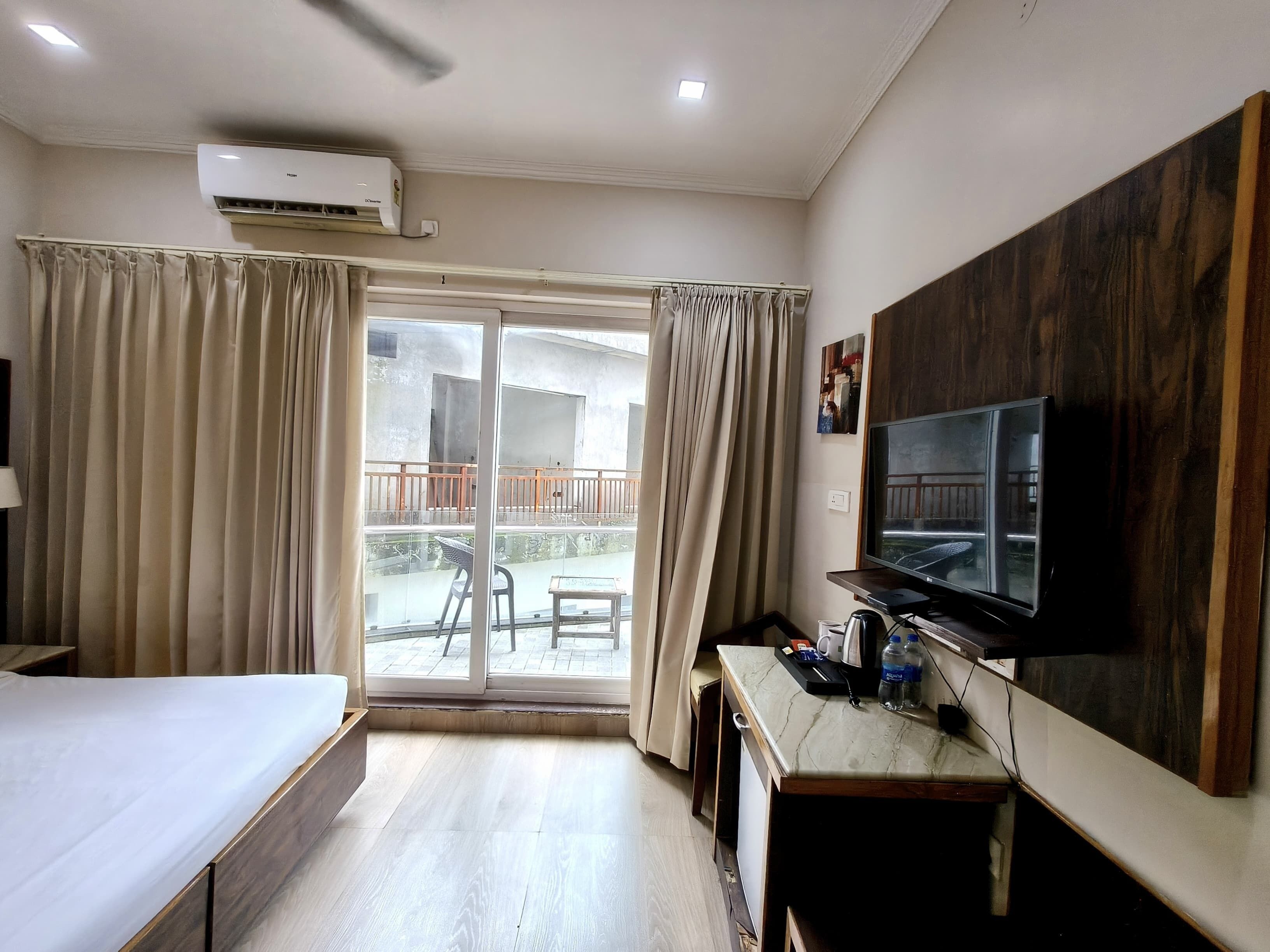 A hotel room with a bed, a desk with a television, a balcony with curtains, and an air conditioning unit.