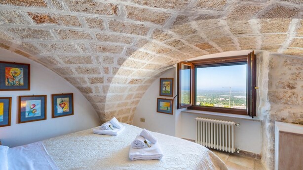A bedroom with a stone ceiling, a bed with white linens, a window with a view, and framed artwork on the wall. - Palazzo Bellavista by Rentbeat (Ostuni)