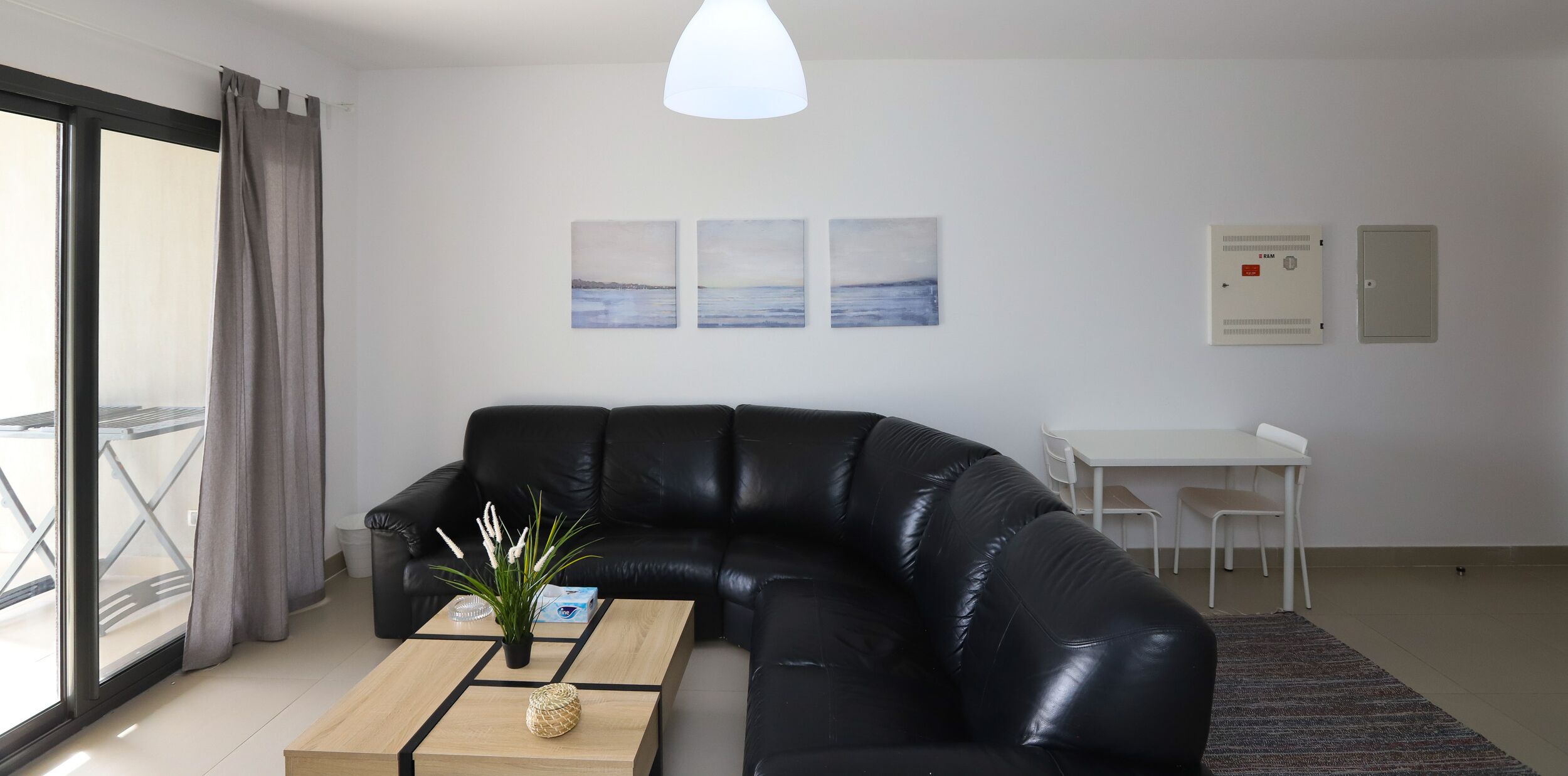 A modern living room with a black leather sofa, a wooden coffee table, and a white pendant light. There is a white table and chairs, a small plant on the coffee table, and a view through a large window with curtains.