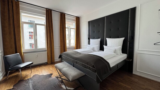 A modern hotel room with a large bed, a bench, a leather rug, and a view of buildings through the windows. - HLH - Luxus Familiensuite mit Terrasse (Freiburg im Breisgau)