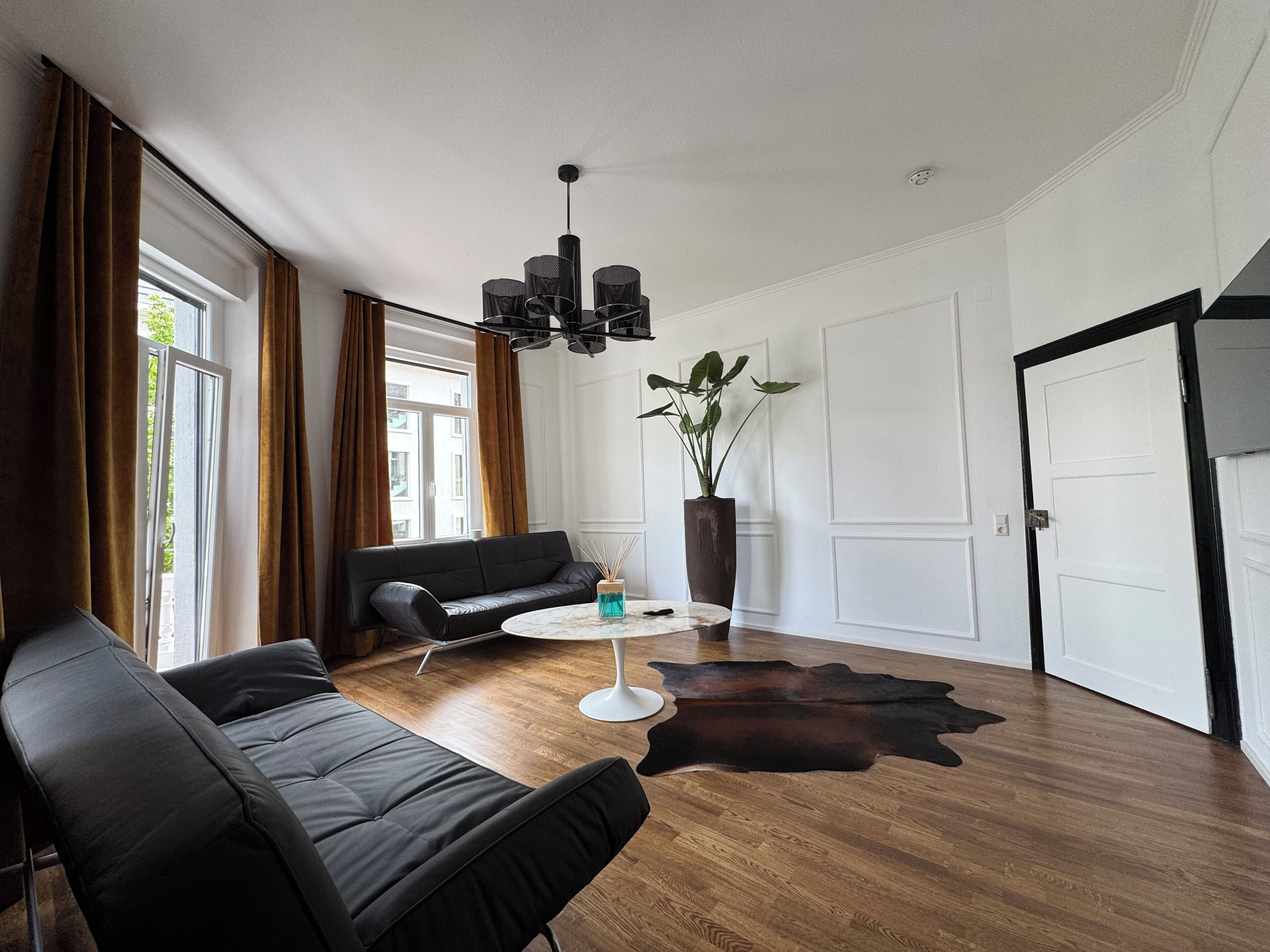 Ein modernes Wohnzimmer mit einem schwarzen Ledersofa, einem runden weißen Tisch, einer großen Topfpflanze und Holzboden.