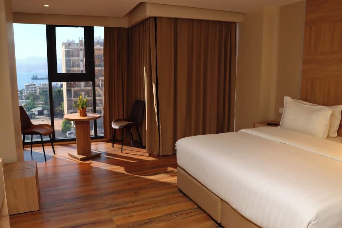 Une chambre d’hôtel avec un grand lit, un bureau avec une chaise, une vue sur la ville grâce à de grandes fenêtres et un balcon.