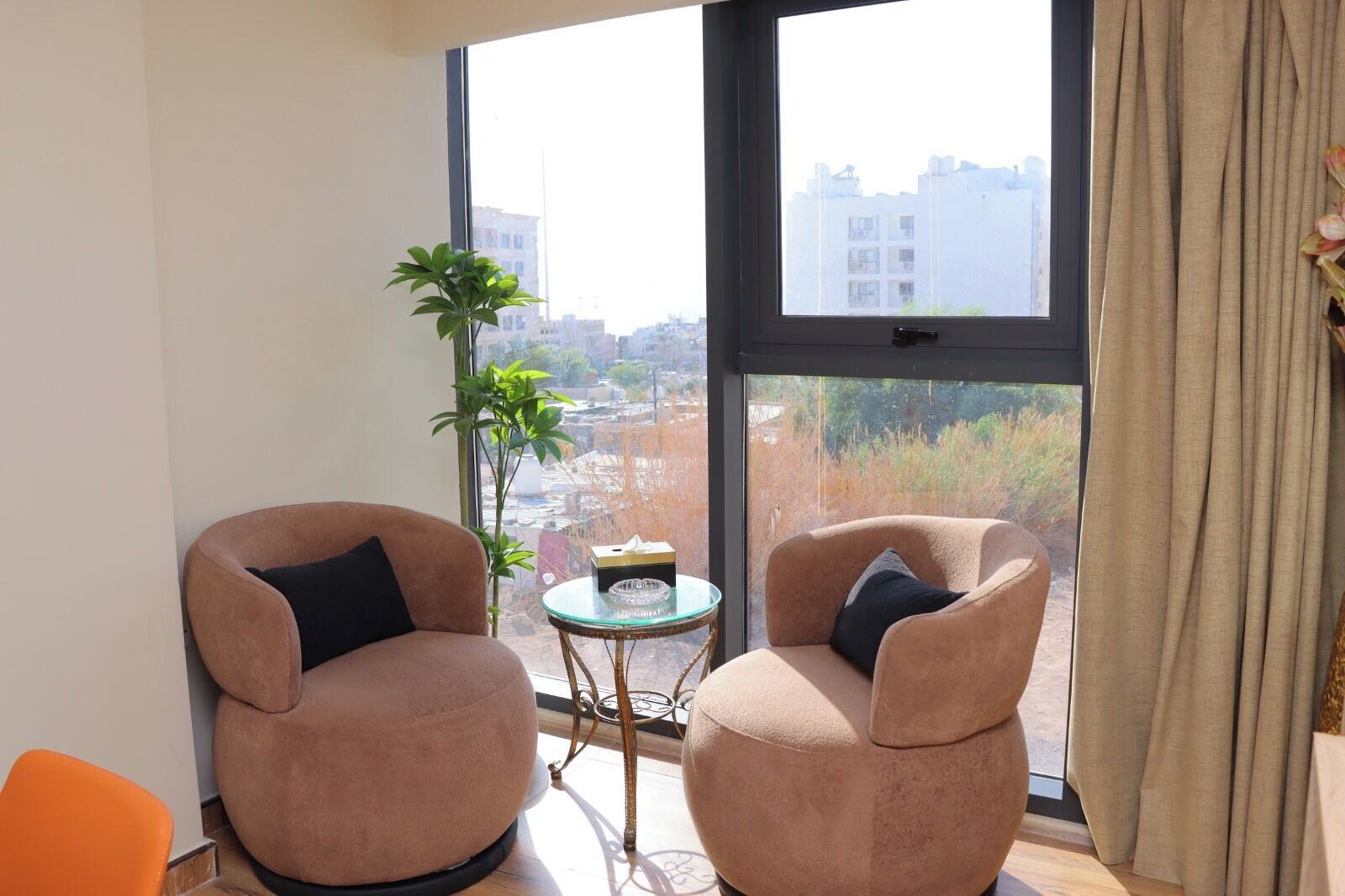 A room with two beige armchairs, a small round table, and a large window offering a city view.