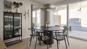 A dining area with a round table, black chairs, a glass cabinet with glassware, a vase with flowers, and a large window with a view of a building and trees.