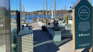An outdoor seating area with tables and chairs, a wooden deck, and a sign for 'Älta Brygga' restaurant.