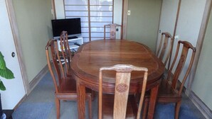 Uma mesa de jantar de madeira tradicional com seis cadeiras, em um chão ladrilhado e com uma janela com persianas.