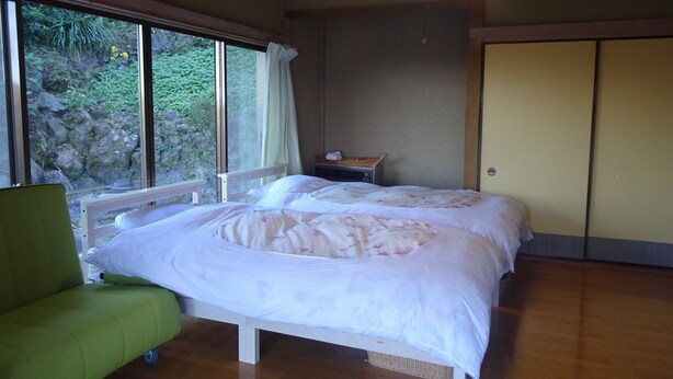 Quarto com duas camas, uma cadeira verde, mesa de cabeceira e janela com vista para o verde.