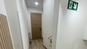 Un pasillo con una puerta de madera al final, un letrero de salida de emergencia en la pared y una pequeña planta decorativa.