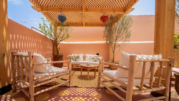 Un área de estar al aire libre con madera, que incluye dos sillas blancas con cojines y una pequeña mesa redonda, rodeada de plantas en macetas y una pérgola de madera.
