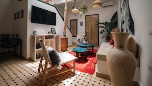 Una habitación con una silla de madera, una mesita pequeña, un jarrón grande y un televisor instalado en la pared.