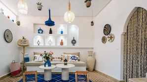 Una habitación con un sofá blanco, dos taburetes de madera, un tapete azul con estampado y varios objetos decorativos en las paredes.