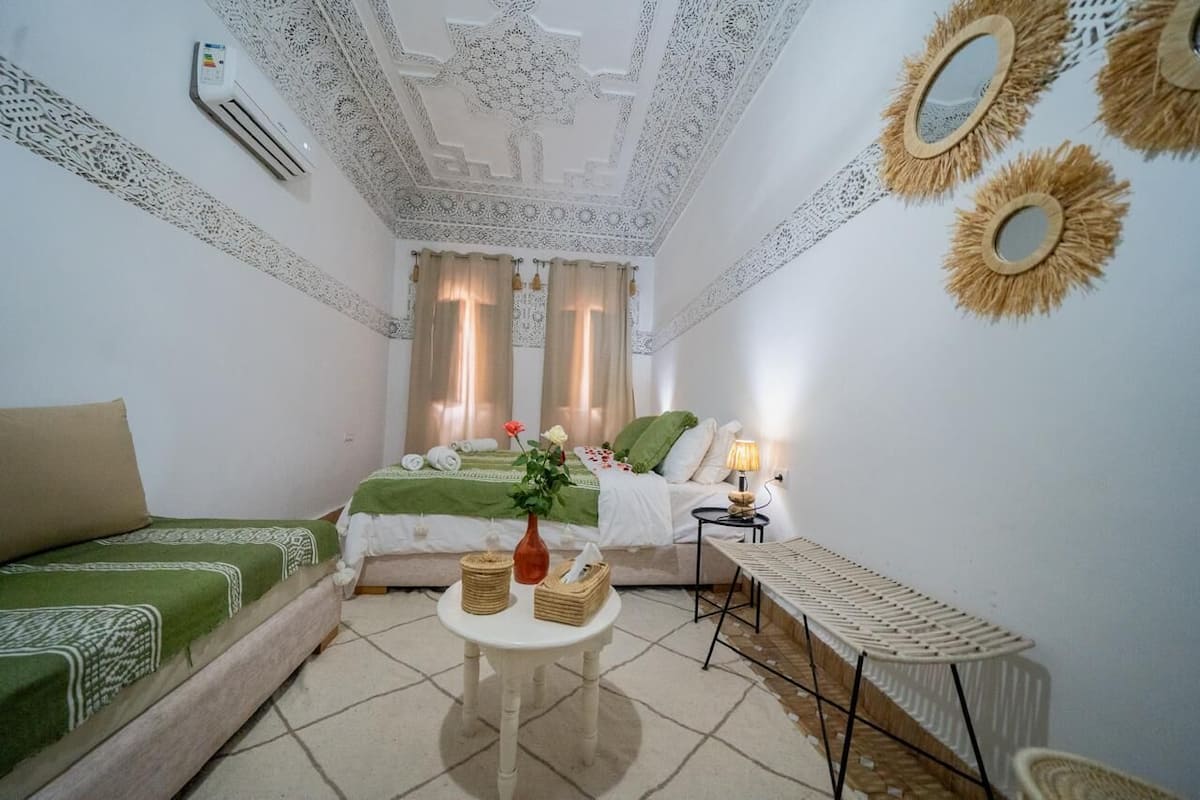 A room with a bed, a green sofa, a small table with baskets, a lamp, and decorative mirrors on the walls.