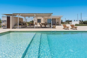 Un área de piscina con zona de estar cubierta, sillas de descanso y una mesa.