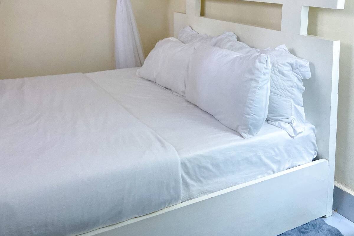 A neatly made bed with white bedding and pillows, a white headboard, and sheer white curtains.
