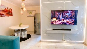 En moderne stue med et fladskærms-TV monteret på en hvid marmorvæg, et spiseområde med et rundt bord og stole, og et køkken med et køleskab.