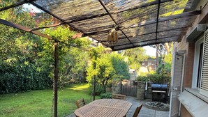 Een overdekt terras met een houten tafel en stoelen, omgeven door weelderige begroeiing en een stenen muur.