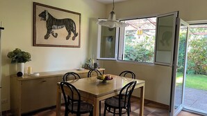 Een eetgedeelte met een houten tafel, vier stoelen, een vaas met groene planten, een lijst met een tijgers afbeelding en een raam met uitzicht op groen.