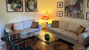 Een woonkamer met twee banken, een glazen salontafel en een lamp. Er hangen afbeeldingen in lijsten aan de muur.