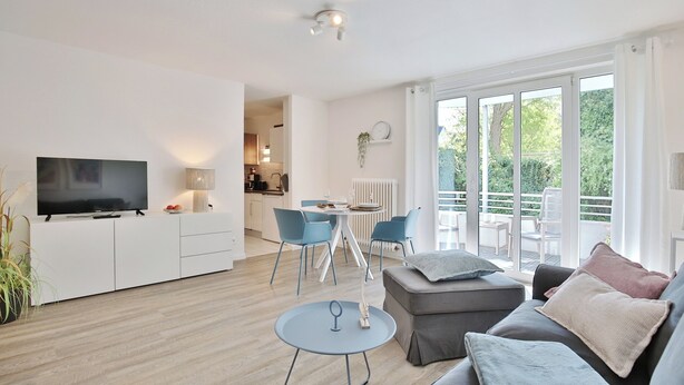 A modern living room with a flat-screen TV, a dining area with blue chairs, a grey sofa with cushions, and large windows offering a view of the outdoors. - Vacation home Bergstraße 62 Apartment 5 Tide am Park (Timmendorfer Strand)