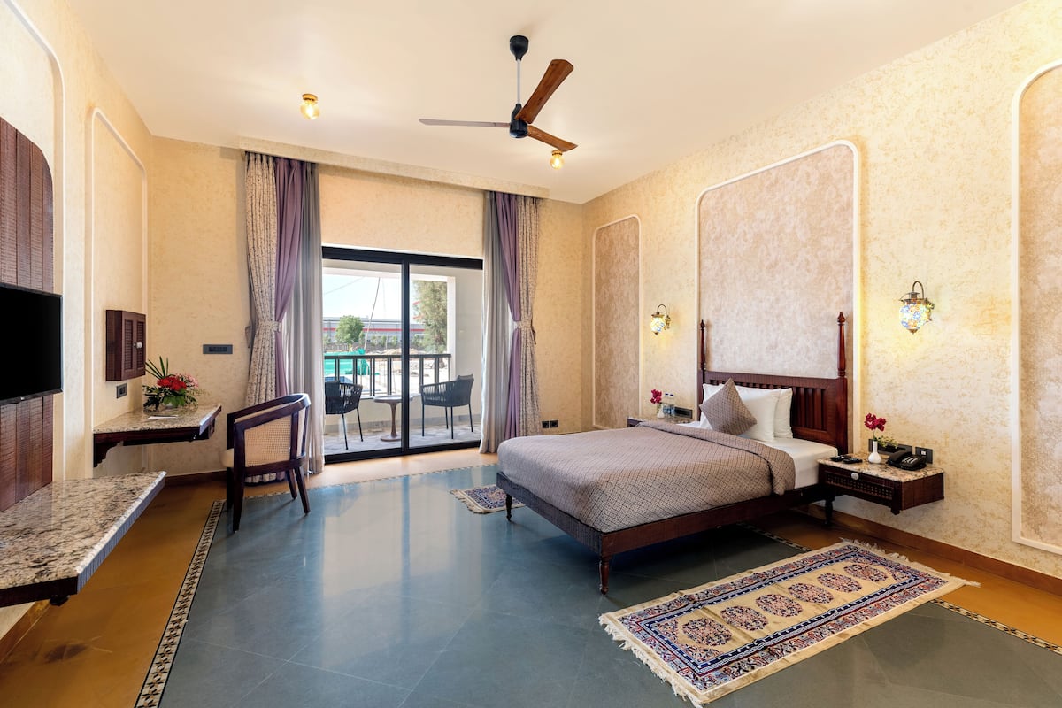 A hotel room with a bed, a desk, a chair, a TV, a ceiling fan, a large window with curtains, and a view of the outside.