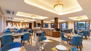 A spacious dining area with high ceilings, chandeliers, and a central bar. Tables are set with blue chairs and white tablecloths. - Chavri Resort – A Luxury Stay In Jawai (Bali)