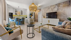 Een moderne woonkamer met een bank, salontafel en stoelen. Achtergrond toont een keuken met witte kasten en een eethoek met blauwe stoelen.
