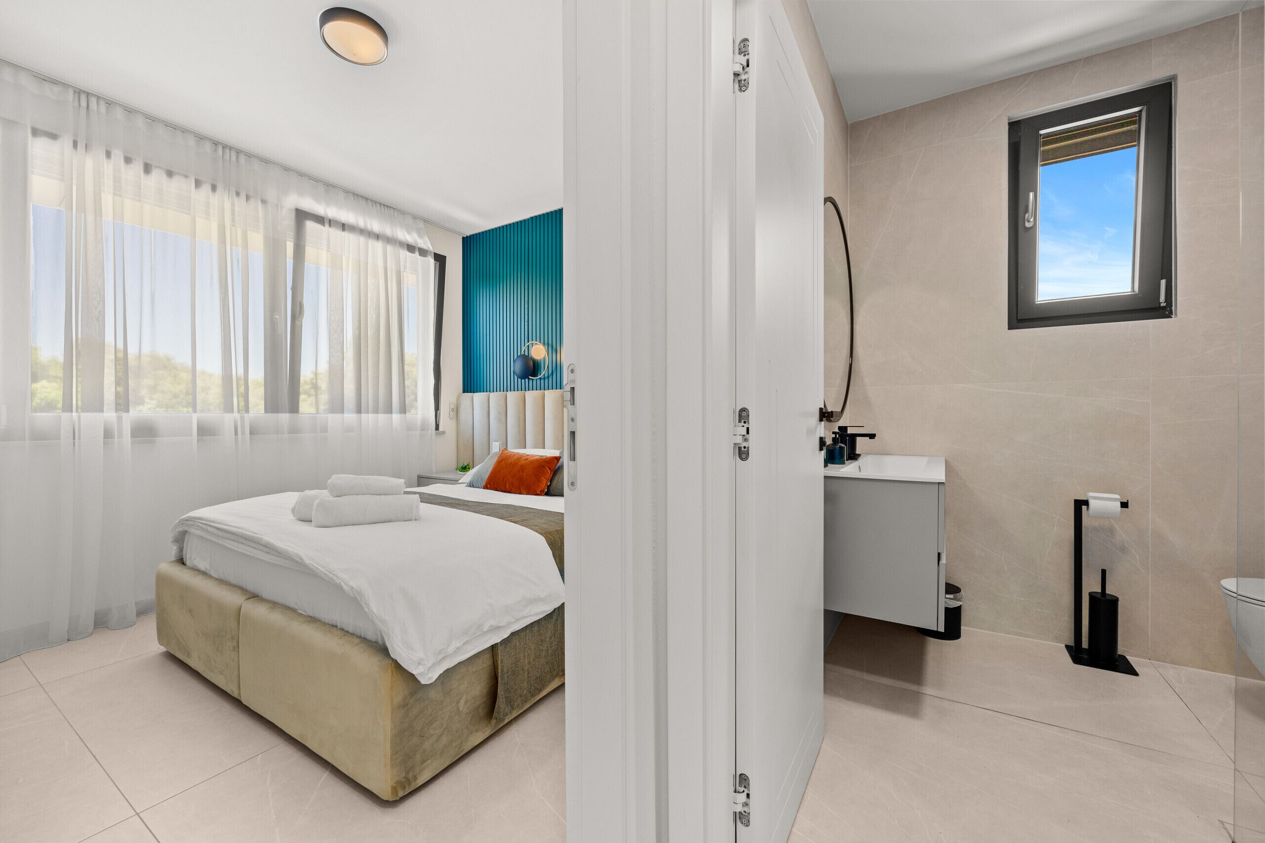 A modern hotel room with a bed, a bathroom with a sink and mirror, and a window with a view.