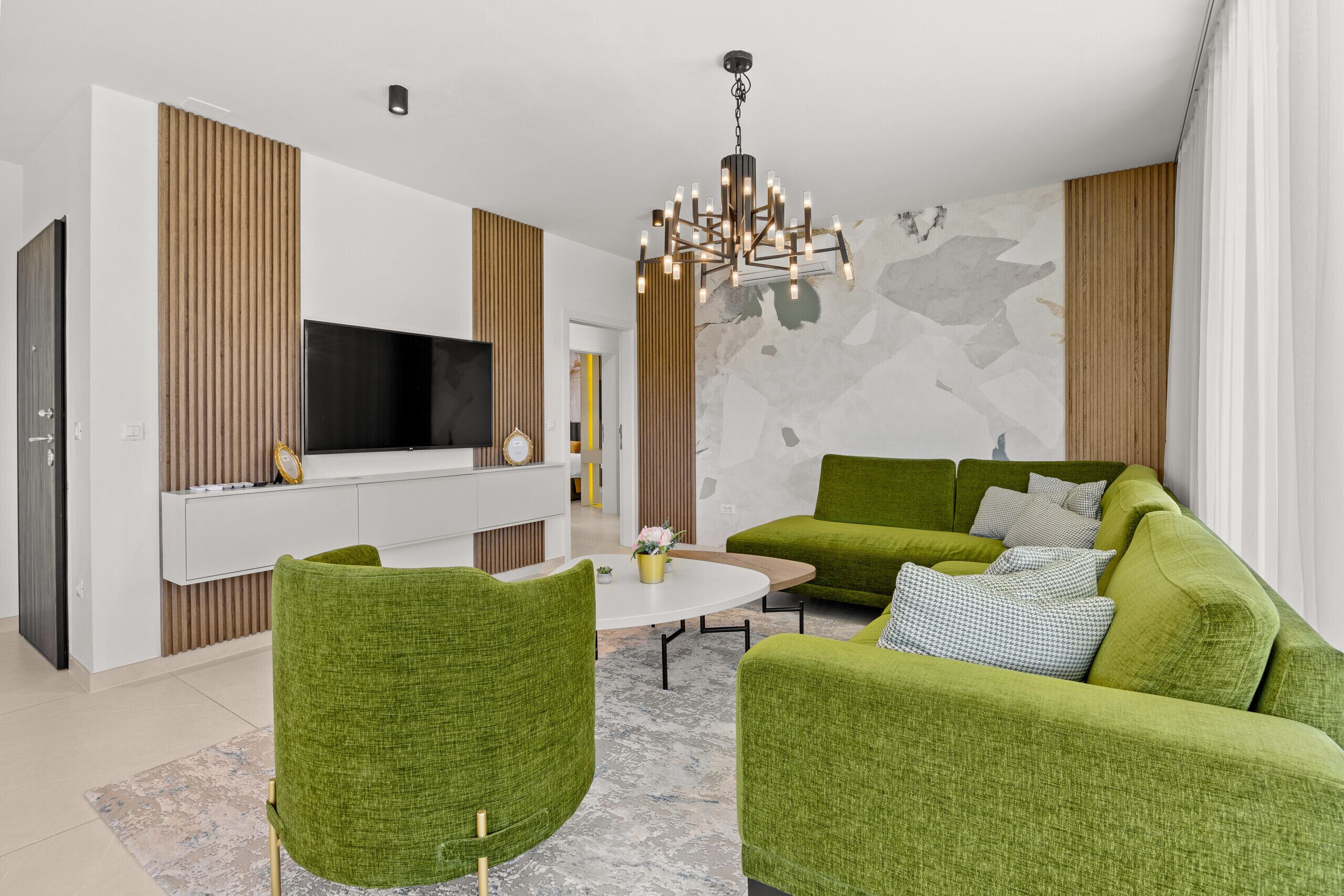 Ein modernes Wohnzimmer mit grün gepolsterten Möbeln, einem Flachbildfernseher und einem dekorativen Kronleuchter.