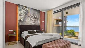 A modern bedroom with a large bed, a tufted ottoman, a nightstand, and a large window with a view of the sea. - The Riviera Three - Luxury Waterfront Apartment (Zadar)