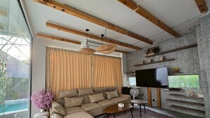 A modern living room with a sectional sofa, a flat-screen TV mounted on a textured wall, and a large window with curtains. - The Ten Poolvilla Huahin (Hin Lek Fai)