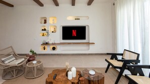 Een moderne woonkamer met een flatscreen televisie aan de muur, een houten plank met decoratieve objecten, rieten meubels en een groot raam met gordijnen.