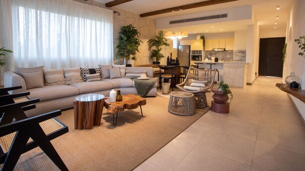 Een moderne woonkamer met een bank, salontafel en stoelen, een keuken op de achtergrond en een groot raam met gordijnen.