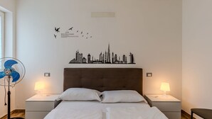 A hotel room with two beds, a city skyline wall art, two bedside lamps, a standing fan, and a wooden floor.