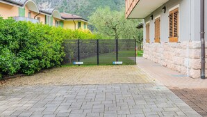 A paved courtyard with a black metal fence, a paved pathway, and a building with stone walls and wooden shutters.