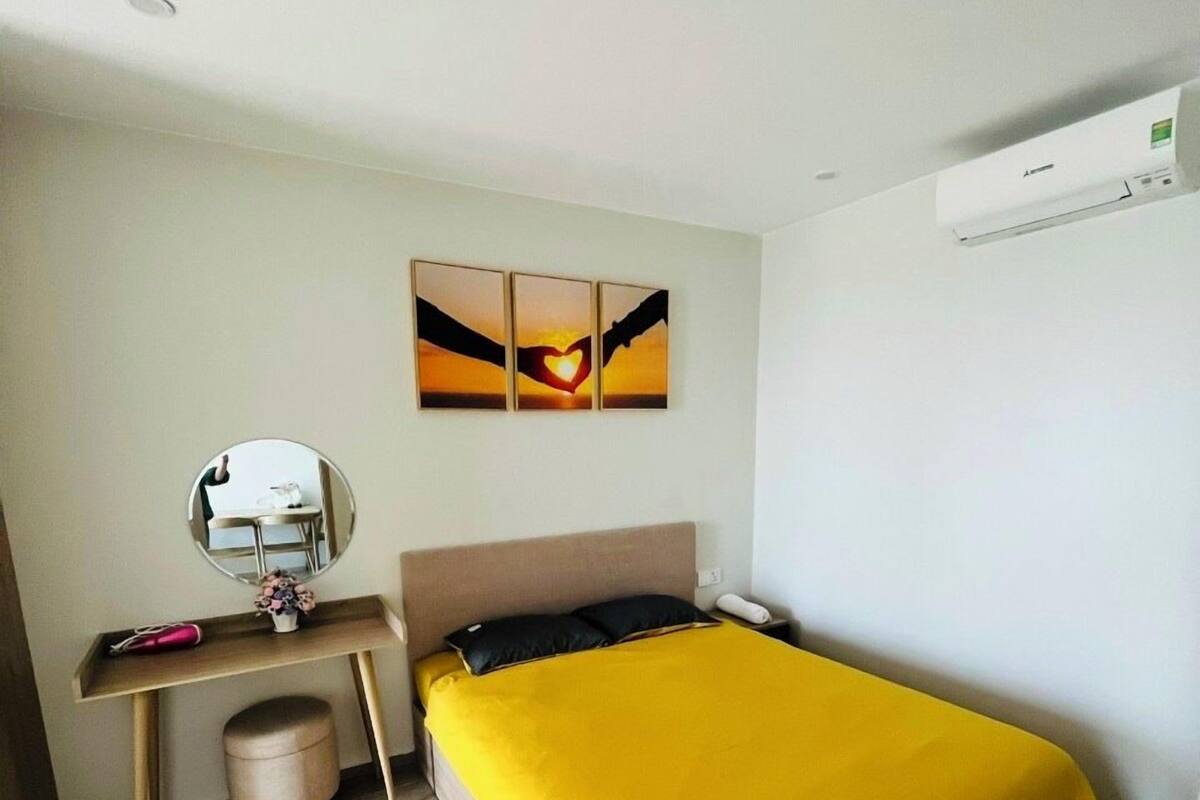 A small, well-lit bedroom with a bed, bedside table, and a mirror. There is a wall-mounted air conditioner and a painting above the bed.