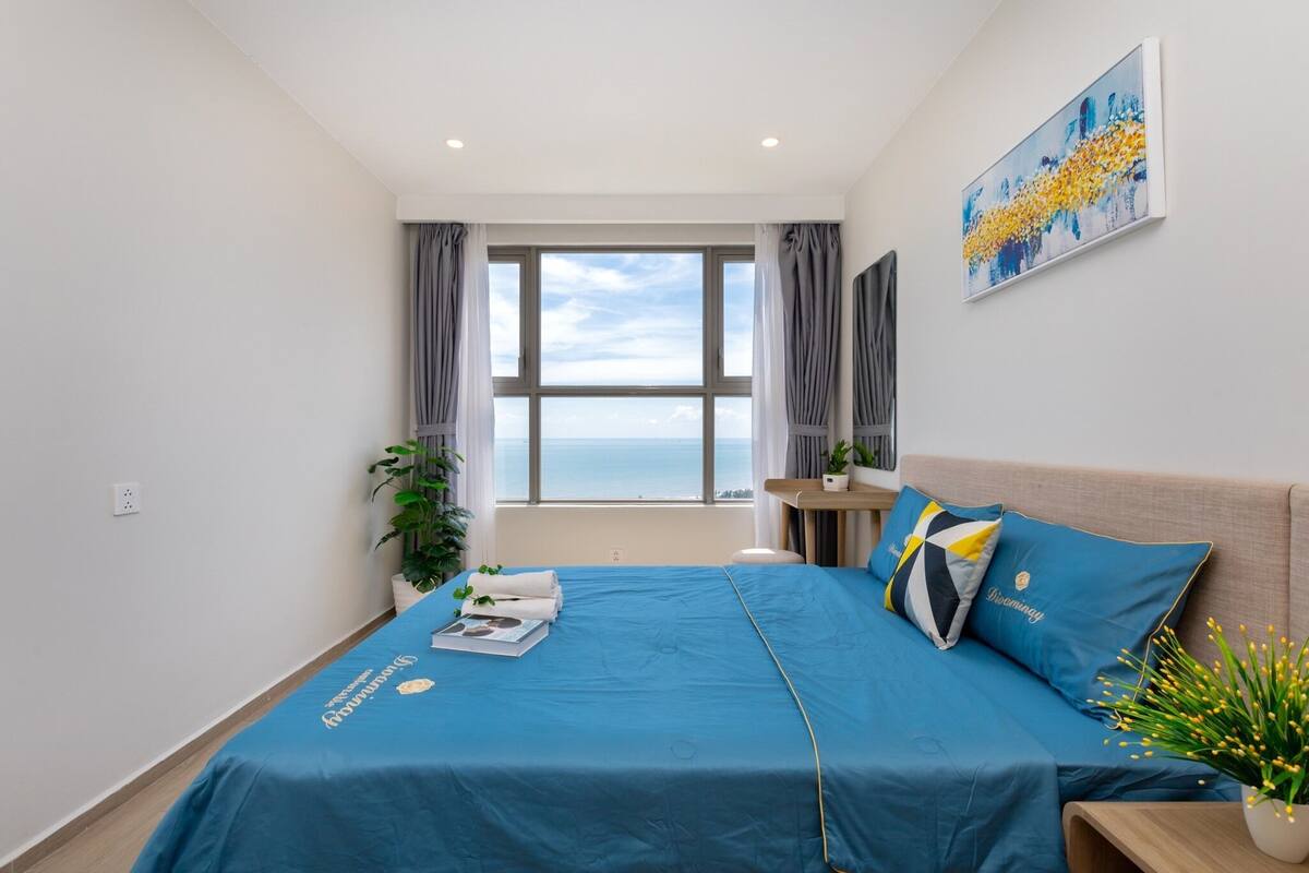 A modern hotel room with a large bed, a view of the sea, a small desk, and a potted plant.