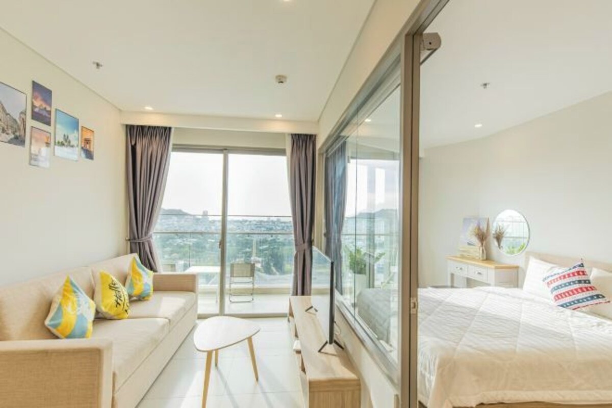 A modern hotel room with a large bed, a sofa, a small table, and a balcony with curtains.
