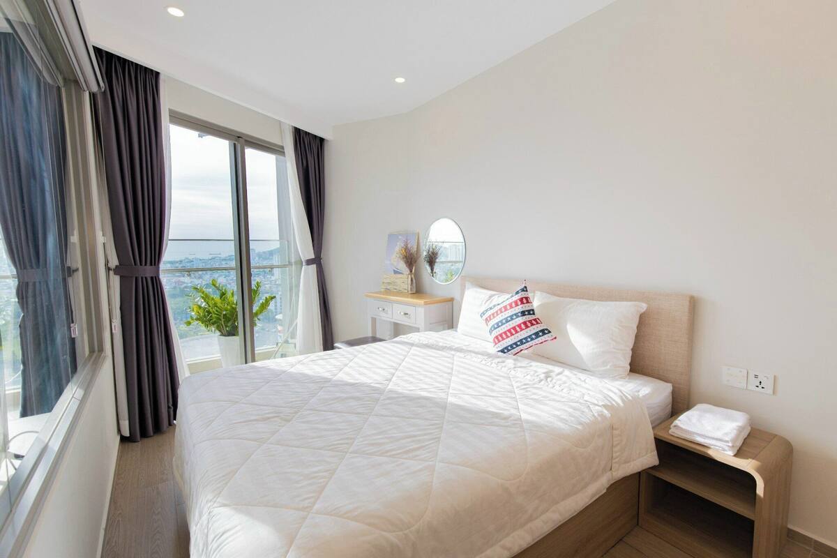 A hotel room with a large bed, a bedside table, a nightstand, a window with curtains, and a view of the city.