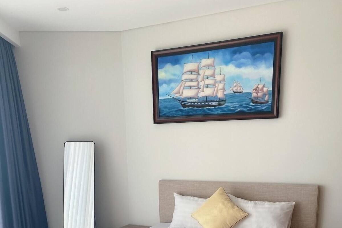 A neatly made bed with a quilted white duvet, a beige headboard, and a single yellow pillow. A framed painting of ships on the sea hangs above the bed.