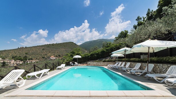 A swimming pool with white lounge chairs and umbrellas, surrounded by stone walls and a view of hills.