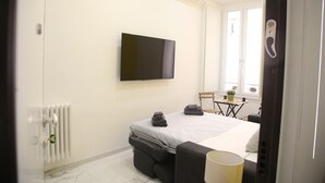A hotel room with a bed, a TV mounted on the wall, a radiator, a small table with a plant, and a window.