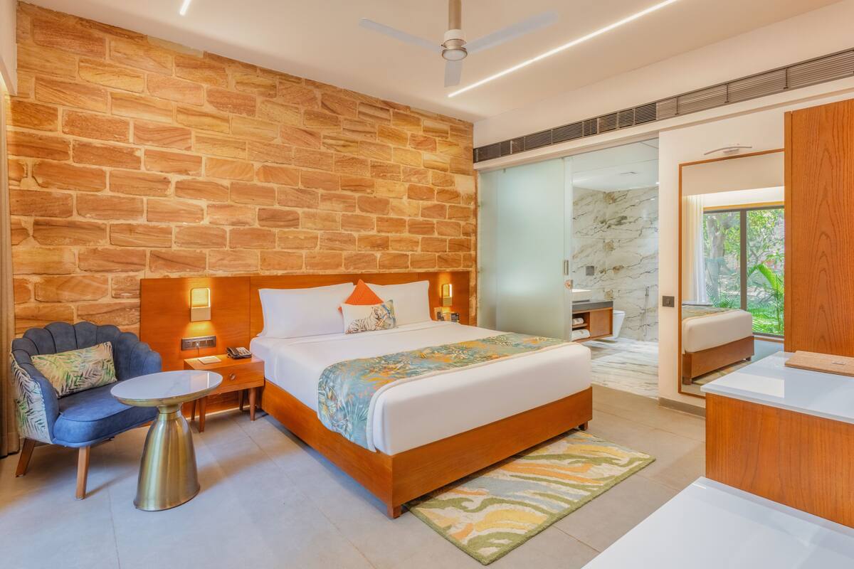A modern bedroom with a stone wall, a bed with a wooden headboard, a nightstand, a chair, a desk, and a view of the outdoors.