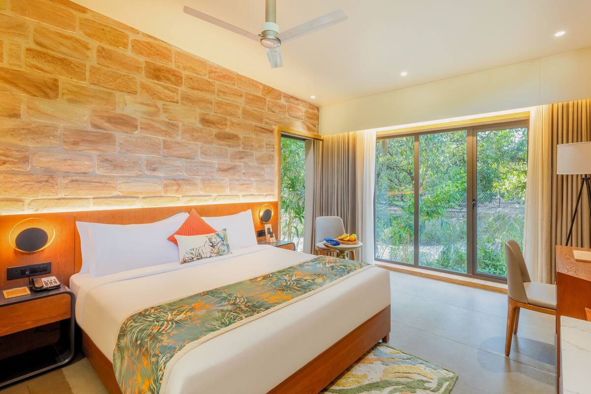 A hotel room with a large bed, a desk, a chair, a ceiling fan, and a view of greenery outside.