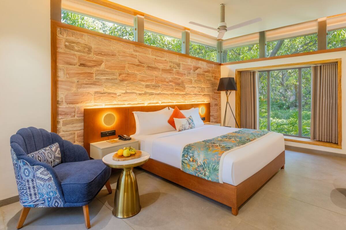 A modern hotel room with a stone wall, a large bed, a round table, and a view of greenery.