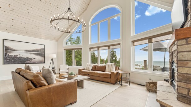 A spacious living room with a brown leather sofa, a coffee table, and a stone fireplace. Large windows offer a view of the outdoors.