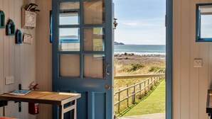 Ein Zimmer mit einem Holztisch, einer blauen Tür, die zum Strand führt, und einem kleinen Fenster mit Meerblick.