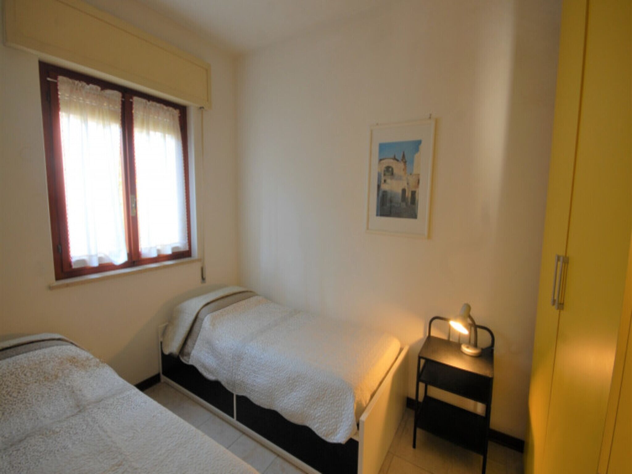 Una stanza piccola e semplice con due letti, un tavolino con una lampada, una finestra con tende e un quadro appeso al muro.