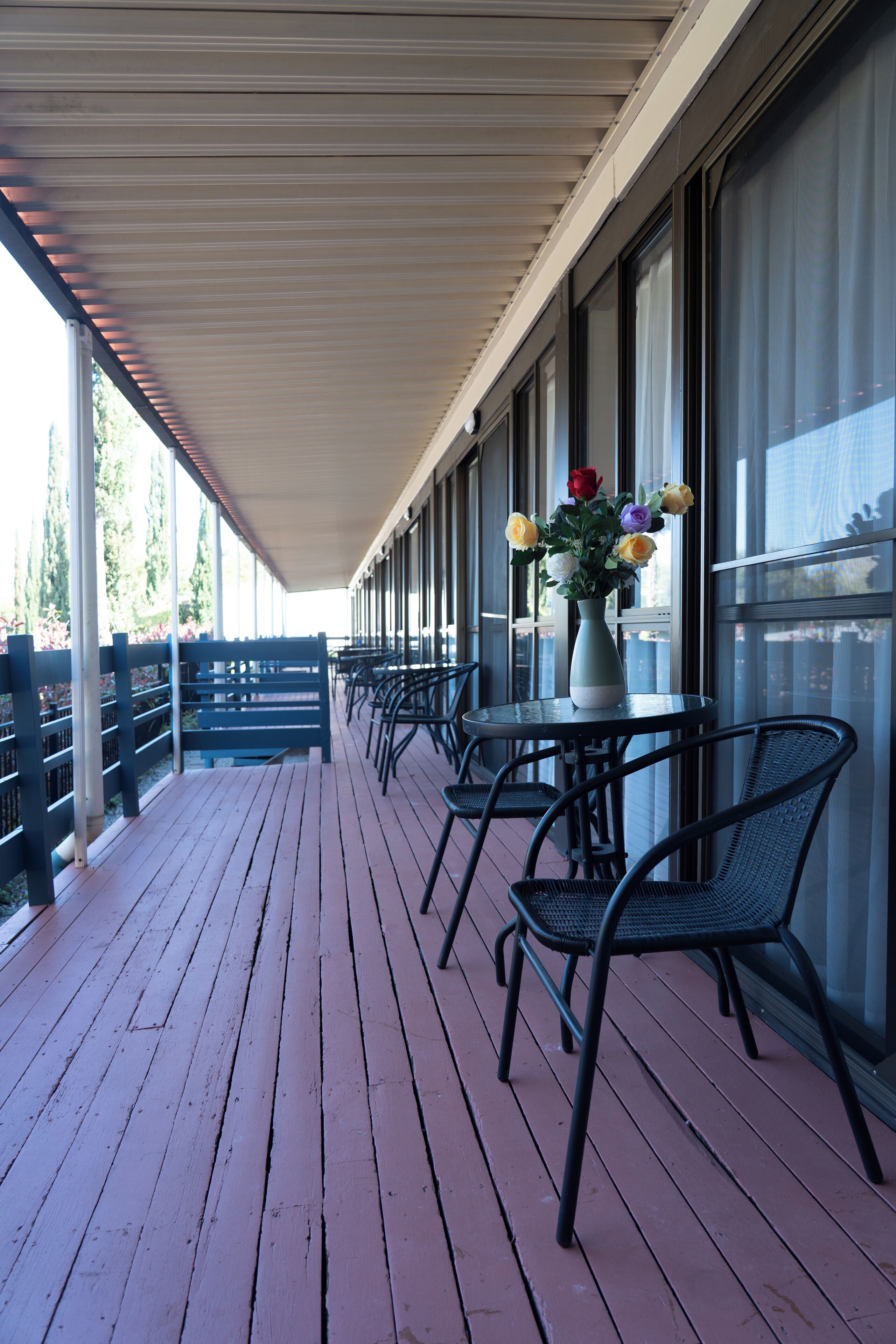 A covered outdoor seating area with tables and chairs, a vase with flowers, and a view through large windows.